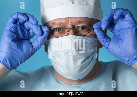 Ein Arzt in medizinischer Kleidung: Eine Mütze, Handschuhe, Morgenmantel und Maske zieht aus, zieht an oder stellt seine Brille ein. Nahaufnahme, schauen Sie sich die Kamera an Stockfoto
