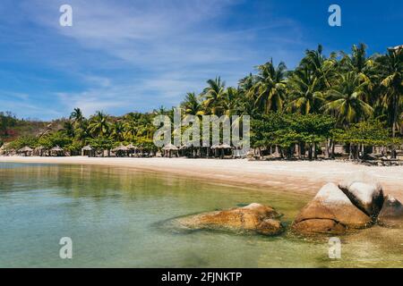 Tropisches Urlaubskonzept. Leerer Sandstrand mit ruhigem Meer, mit klarem blauen Himmel. Kokospalmen, größere Steinbrocken im Hintergrund Stockfoto