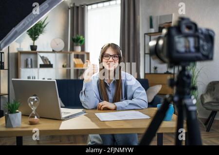 Kaukasische junge Frau mit Headset und Brille zeigt Daumen nach oben, während sie ein Video mit der Kamera filmt. Weibliche Coach Erstellung Online-Tutorial während zu Hause bleiben. Stockfoto