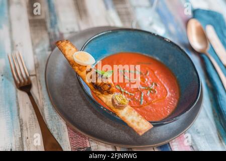 Kalte Tomatensuppe mit Olivenöl, Kräutern in Stückchen und Arten, die auf einem rustikalen Holztisch neben Besteck verzehrt werden können. Stockfoto