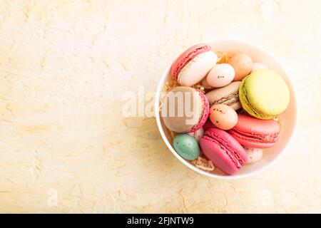 Bunte Makronen und Schokoladeneier in Keramikschale auf beigefarbenem Beton-Hintergrund. Draufsicht, Kopierraum, Stillleben. Frühstück, Morgen, Konzept. Stockfoto