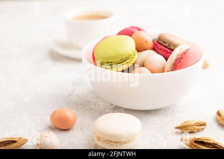 Bunte Makronen und Schokoladeneier in Keramikschale, Tasse Kaffee auf grauem Betongrund. Seitenansicht, Nahaufnahme, selektiver Fokus, Stillleben Stockfoto
