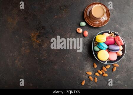 Bunte Makronen und Schokoladeneier in Keramikschale, Tasse Kaffee auf schwarzem Betongrund. Draufsicht, flach liegend, Kopierraum, Stillleben. Bre Stockfoto