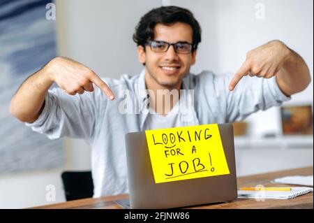 Verschwommener Arbeitsloser positiver Hispanic mit Brille sitzt an seinem Schreibtisch, zeigt mit den Fingern auf ein Schild auf einem Laptop mit einer Aufschrift auf der Suche nach einem Job, schaut auf die Kamera und lächelt freundlich Stockfoto