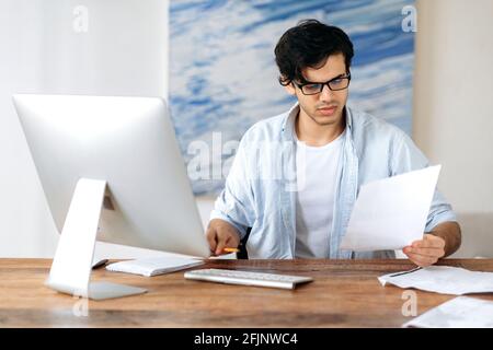 Fokussierter, intelligenter hispanischer Mann mit Brille, Manager, Freiberufler oder Büroangestellter, der am Tisch mit Computer arbeitet, Geschäftsdokumente durchschaut, Finanzbericht analysiert Stockfoto