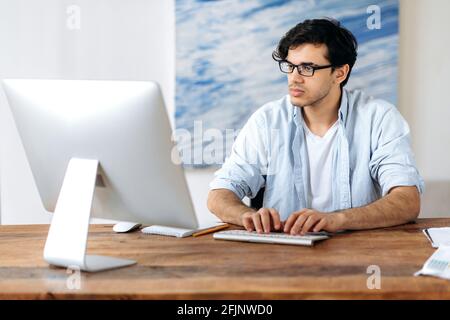 Intelligenter selbstbewusster junger Hispanic-Mann mit Brille, Manager, Geschäftsmann oder Freiberufler, stilvoll gekleidet, am Computer arbeitend, mit einem Kollegen oder Kunden schickend, im Internet surfend, per E-Mail Stockfoto