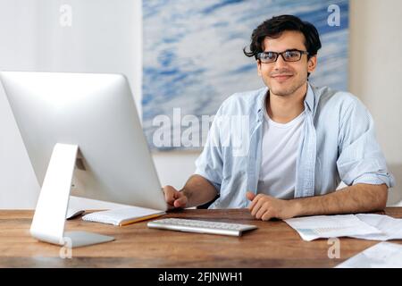 Porträt eines gutaussehenden, smarten, erfolgreichen jungen hispanischen Geschäftsmannes, Managers oder Freiberuflers, in stilvoller Kleidung und Brille, sitzt am Arbeitsplatz, arbeitet am Computer, blickt auf die Kamera und lächelt angenehm Stockfoto