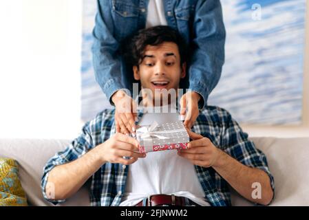 Überrascht glücklicher junger hispanischer Mann, der in legerer Kleidung auf dem Sofa im Wohnzimmer sitzt und von seiner geliebten Freundin, seinem Mädchen hinter ihm, eine schöne Geschenkbox erhält, schaut er erstaunt auf eine Schachtel Stockfoto