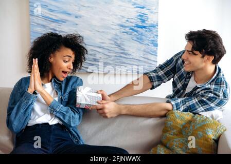Auf der Couch sitzend freudig schockierte junge hübsche afroamerikanische Frau erhält von einem liebenden Latino-Typen eine Geschenkbox für einen Geburtstag oder Beziehungsjubiläum, schaut auf ein Geschenk, klatscht in die Hände Stockfoto