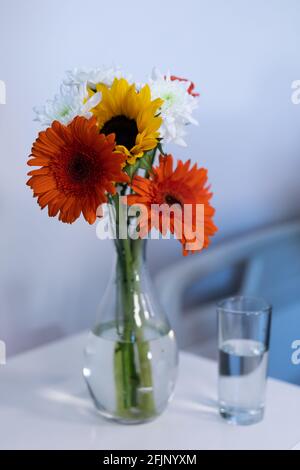 Bunte Gerbera Blumen in einer Vase und ein Glas Wasser auf dem Nachttisch eines Krankenhauspatienten Stockfoto