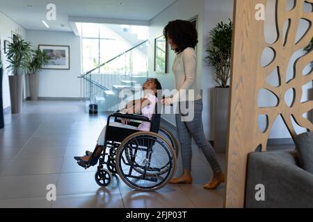 Glückliche Mischling Mutter schiebt lächelnde Tochter im Rollstuhl durch Krankenhausfoyer Stockfoto
