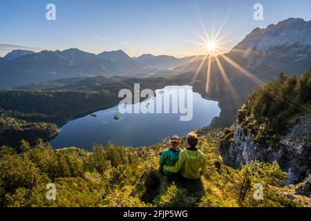 Zwei (Wanderer) blicken bei Sonnenaufgang auf den Eibsee, die Sonne scheint über den bayerischen Voralpen, die rechte Zugspitze, das Wettersteingebirge bei Grainau Stockfoto