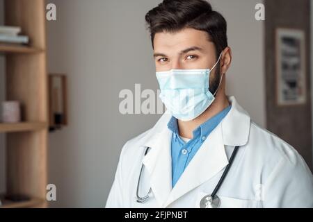 Kaukasischen jungen Arzt im Gesicht Maske weißen medizinischen Mantel mit Stethoskop, Hochformat Stockfoto