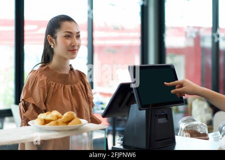 Ein junger asiatischer Kunde bestellt ein Getränk vor der Kasse. Der Kellner erhält auf der anderen Seite Bestellungen mit einem Computermonitor. Morgenstimmung Stockfoto