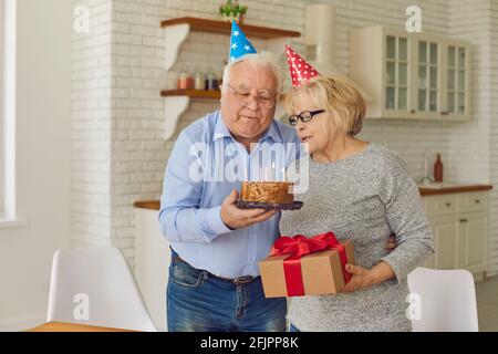 Glückliches reifes Paar in kegelförmigen Hüten, das Kerzen auf einem Kuchen ausbläst. Stockfoto