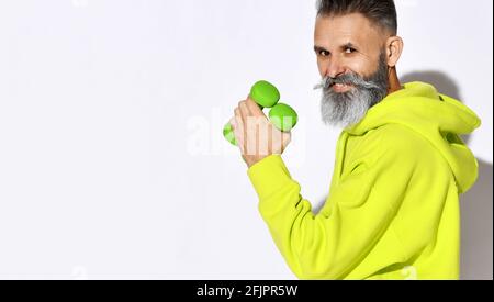 Porträt eines fröhlichen, aktiven alten, brutalen bärtigen Mannes in gelbem Kapuzenpullover, der zwei grüne Hanteln in der Hand hält. Seitenansicht Stockfoto