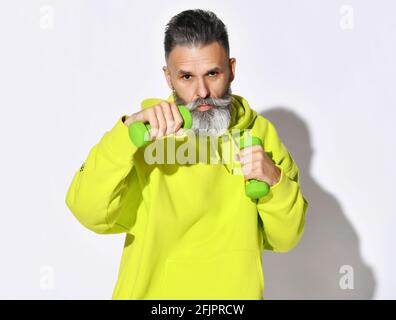 Porträt eines alten brutalen bärtigen Mannes in gelbem Kapuzenpullover Fitness-Training Boxen mit grünen Hanteln in der Hand Stockfoto