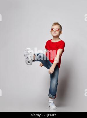 Frolic Kid Boy Teenager in rotem T-Shirt, blauer Jeans, weißen Turnschuhen und Sonnenbrillen steht mit dem Fuß nach oben Stockfoto