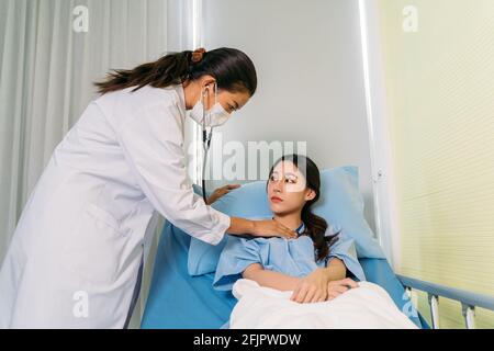 Junger Arzt im Laborkittel mit Gesichtsmaske, die den Herzrhythmusrhythmus überprüft Mit Stethoskop einer kranken asiatischen Patientin, die im Krankenhaus liegt Bett Stockfoto