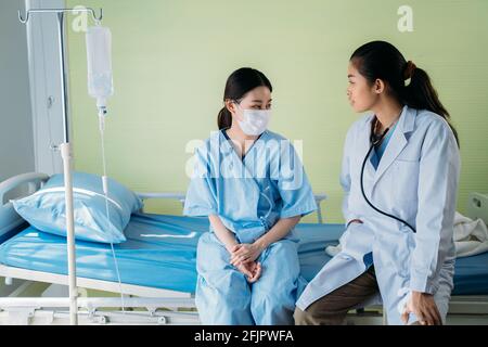Junge asiatische Ärztin mit Patient trägt chirurgische Gesichtsmaske Auf dem Krankenhausbett sitzend und miteinander redeten und redeten Routinemäßige Statusaktualisierung Stockfoto