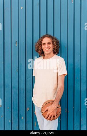 Vertikales Porträt eines jungen kaukasischen Mannes, der lächelnd auf die Kamera schaut und einen Basketball hält. Er hat lange rote Haare und trägt lässige Sommerkleidung. Es lehnt sich an eine blaue Metalltür Stockfoto