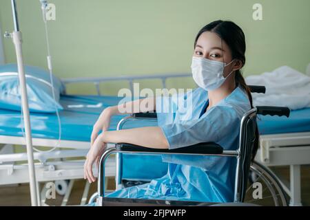 Porträt einer jungen asiatischen behinderten Frau mit traurigen und hoffnungslosen Gefühl, allein im Krankenhaus im Rollstuhl zu sitzen und einen chirurgischen Eingriff zu tragen Gesichtsmaske Stockfoto