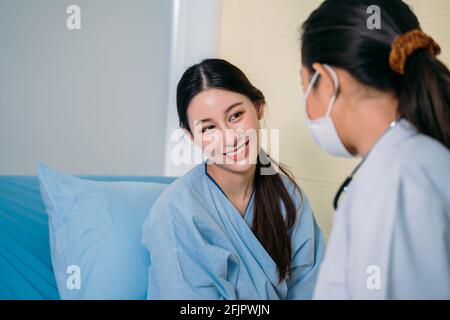 Porträt von schönen jungen asiatischen weiblichen Patienten in Scrubs sprechen Zum Arzt, der aus Sicherheitsgründen eine Gesichtsmaske trägt, während er auf dem Arzt sitzt Krankenhausbett während der Konsultation Stockfoto