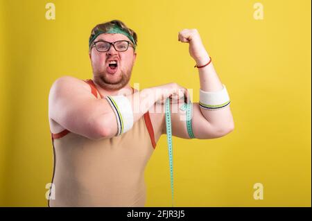 Ein lustiger, fröhlicher, fetter Mann in Sportkleidung mißt seine Arme. Übergewichtiger Mann geht in den Sport. Gelber Hintergrund isoliert Stockfoto