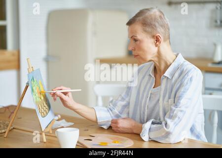 Kreatives Hobby. Seitenansicht einer konzentrierten reifen Frau, die zu Hause ein Bild malt, während sie am Holztisch in der Küche sitzt, selektiver Fokus auf Frau. Senior People und Kunstkonzept Stockfoto