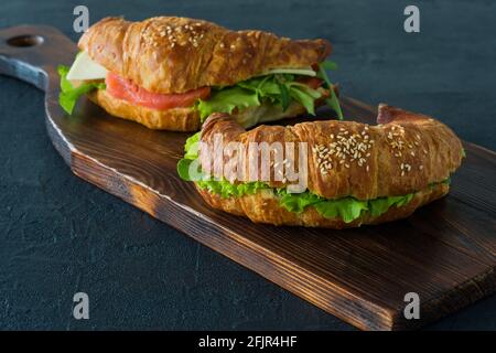 Croissant-Sandwiches mit gesalzenem Lachs auf einem Schreibtisch, serviert mit frischen Salatblättern, Rucola und Gemüse auf schwarzem Hintergrund. Stockfoto