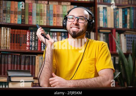 Ein kahler Mann mit Brille und Bart, der ein Hörbuch hört oder Musik mit Kopfhörern hört. Bücherregale im Hintergrund. Konzept der Online-Entertainer Stockfoto