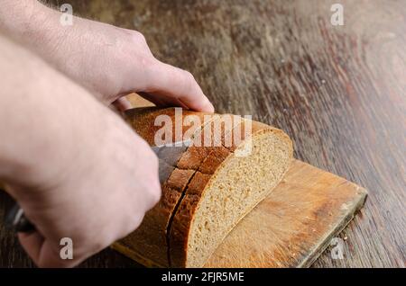 Hände schneiden Brot auf einem Holzschneidebrett. Ein gewachsener Mann schneidet mit einem großen Messer einen Laib Roggenbrot in Scheiben. Nahaufnahme. Selektiver Fokus. Stockfoto