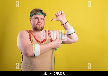 Ein lustiger, fröhlicher, fetter Mann in Sportkleidung mißt seine Arme. Übergewichtiger Mann geht in den Sport. Gelber Hintergrund isoliert Stockfoto