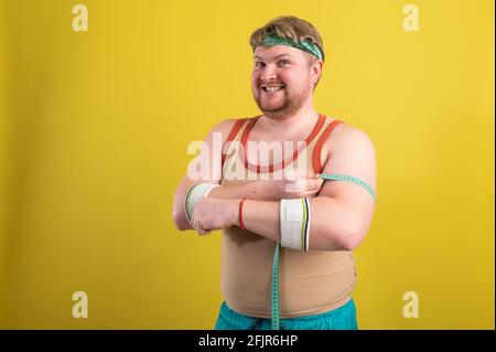 Ein lustiger, fröhlicher, fetter Mann in Sportkleidung mißt seine Arme. Übergewichtiger Mann geht in den Sport. Gelber Hintergrund isoliert Stockfoto