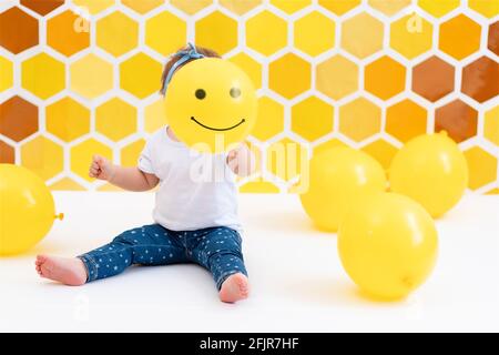 Ein Kleinkind Mädchen sitzt auf einem weißen Boden mit gelben Ballons., hält lächelnde Ballon in der Nähe des Gesichts. Im Hintergrund sind gelbe Waben zu sehen. Die CO Stockfoto