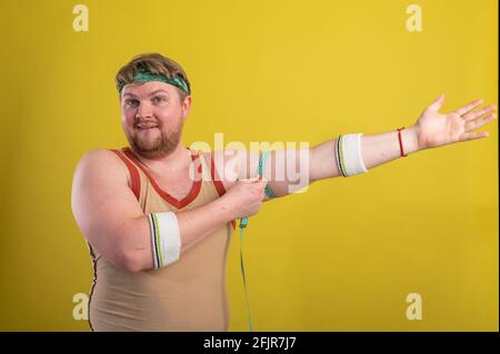 Ein lustiger, fröhlicher, fetter Mann in Sportkleidung mißt seine Arme. Übergewichtiger Mann geht in den Sport. Gelber Hintergrund isoliert Stockfoto