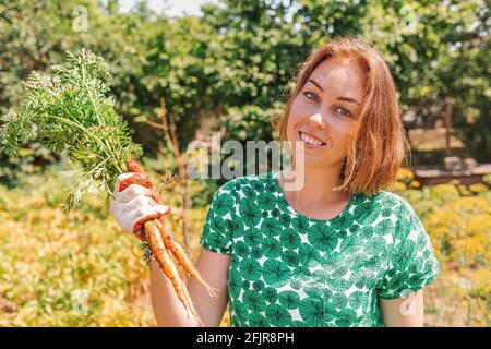 Eine kaukasische junge Frau lächelt und hält ein paar frisch gepflückte Karotten. Vegetation im Hintergrund.Konzept der Ernte und Gartenarbeit. Stockfoto