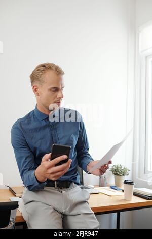 Nachdenklicher, seriöser junger Geschäftsmann mit Smartphone in der Hand, auf dem er sitzt Seinen Tisch und Lesebericht oder gedruckte E-Mail Stockfoto