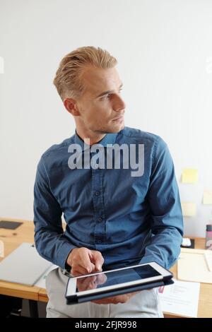 Portrait des jungen gutaussehenden Unternehmers mit Anwendung auf Tablet-Computer Und durch das Bürofenster schauen Stockfoto