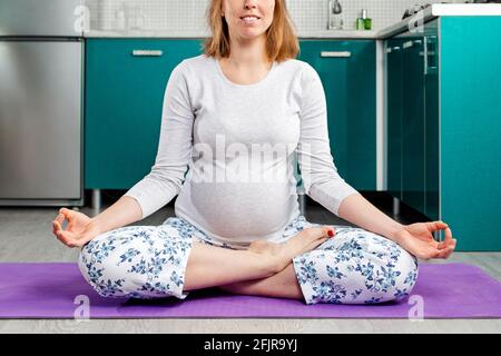 Eine schwanger kaukasische Frau macht Yoga und meditiert mit gekreuzten Beinen auf einer Matte. Die Küche ist im Hintergrund. Konzept von Gesundheit und Yoga für Schwangere wo Stockfoto