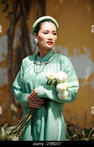 Porträt der schönen jungen vietnamesischen Frau in traditioneller Kleidung und Kopfbedeckung mit Lotusblumen und Blick weg Stockfoto