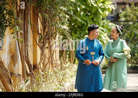 Glückliches junges asiatisches Paar in traditionellen Kleidern und Kopfteilen Lachen und reden, wenn man am Sommertag draußen läuft Stockfoto