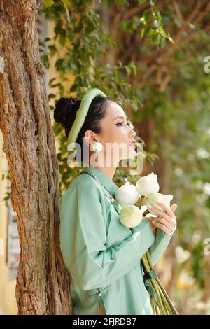 Nachdenklich schöne junge vietnamesische Frau in traditionellem Kleid mit Strauß Von Blumen und auf Baum gelehnt Stockfoto