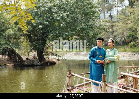 Frisch verheiratetes asiatisches Paar in traditionellen Kostümen, das am Teich steht Im Stadtpark und Blick auf das Wasser Stockfoto