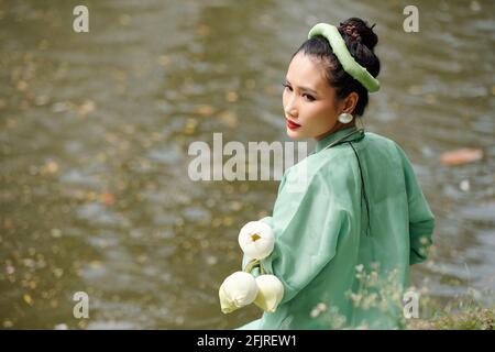 Ernst attraktive junge vietnamesische Frau steht am Wasser mit Blumenstrauß Von Blumen und Umkehren Stockfoto