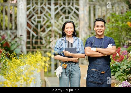 Porträt lächelnder junger vietnamesischer Arbeiter, die im Gewächshaus stehen, die Arme kreuzen und die Kamera betrachten Stockfoto