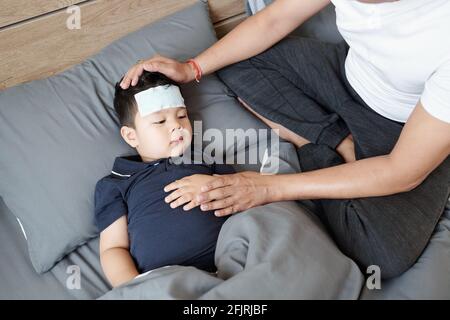 Vater sitzt neben seinem kranken Sohn, der sich im Bett ausruht Mit kühlenden Gelen auf der Stirn, die an Fieber leidet Stockfoto