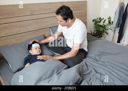 Vater kümmert sich um sein kleines Kind mit Fieber liegen Im Bett zu Hause mit kühlenden Flecken auf der Stirn Stockfoto
