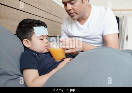 Fürsorglicher Vater, der ihm ein Glas frischen Orangensaft gab Krankes kleines Kind liegt im Bett mit Kühlfleck auf Stirn Stockfoto