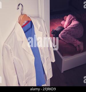 Müder Arzt schläft zu Hause auf einem Bett mit einer Katze, weiße medizinische Uniform auf einem Kleiderbügel im Schlafzimmer Stockfoto